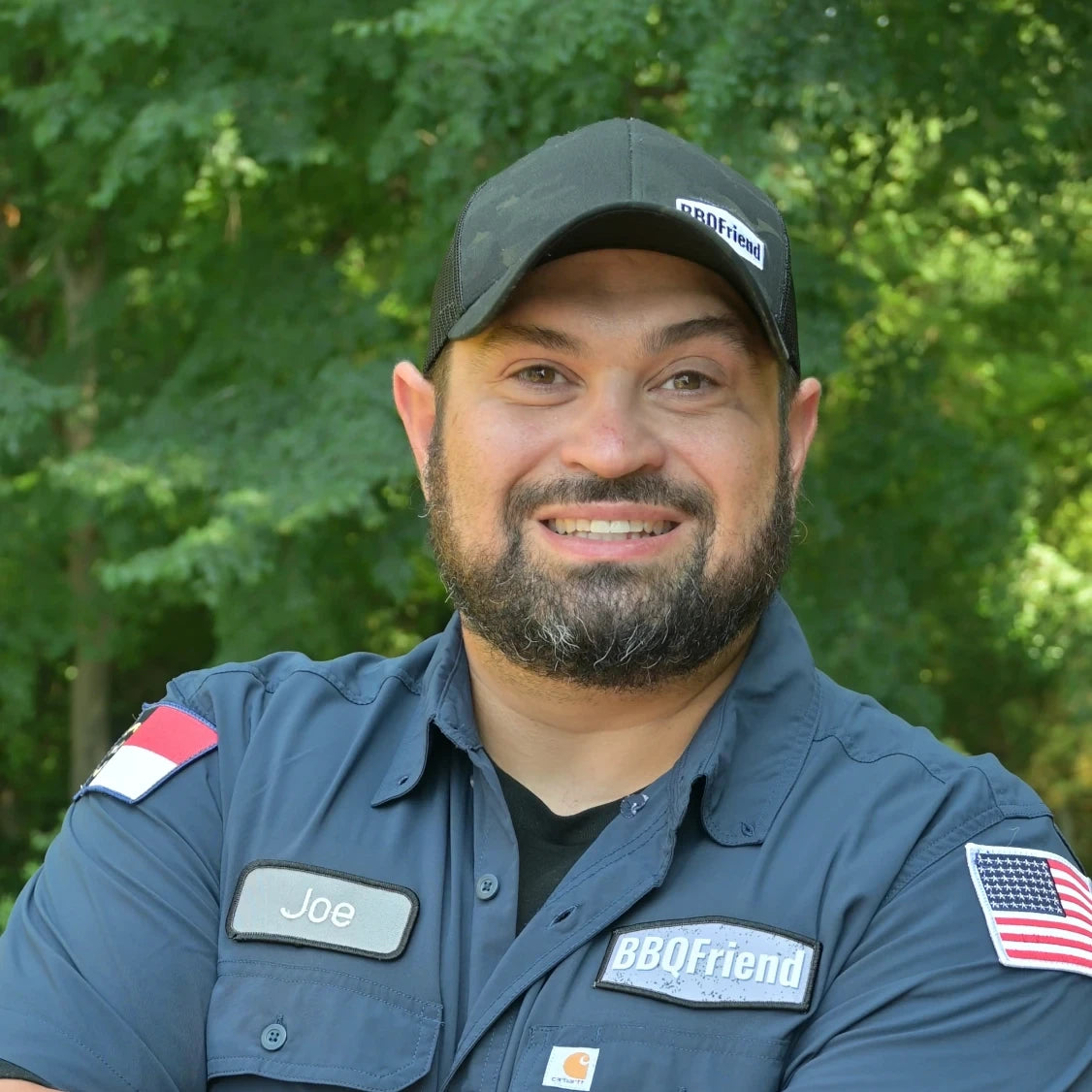 Joe DeApice stands smiling with arms crossed. He is wearing a blue BBQFriend shirt with an American flag on the sleeve.