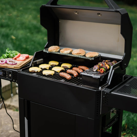 An AutoIgnite grill with the lid open to show 6 cheeseburgers, 4 sausages, 4 hamburger buns and 2 skewers of vegetables all cooking at the same time. The left side shelf holds a wood cutting board with lettuce leaves and onion and tomato slices.