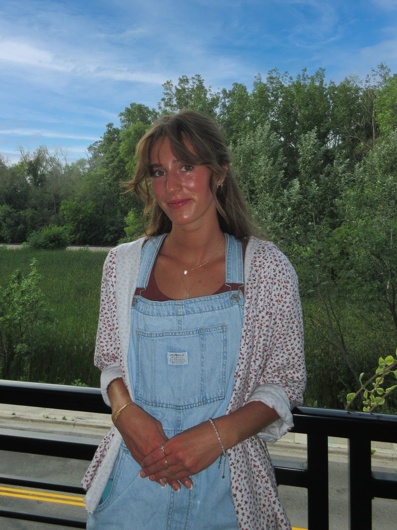 Lauren Sephton wearing faded blue overalls and a light colored shirt leans against a railing in front a wooded area