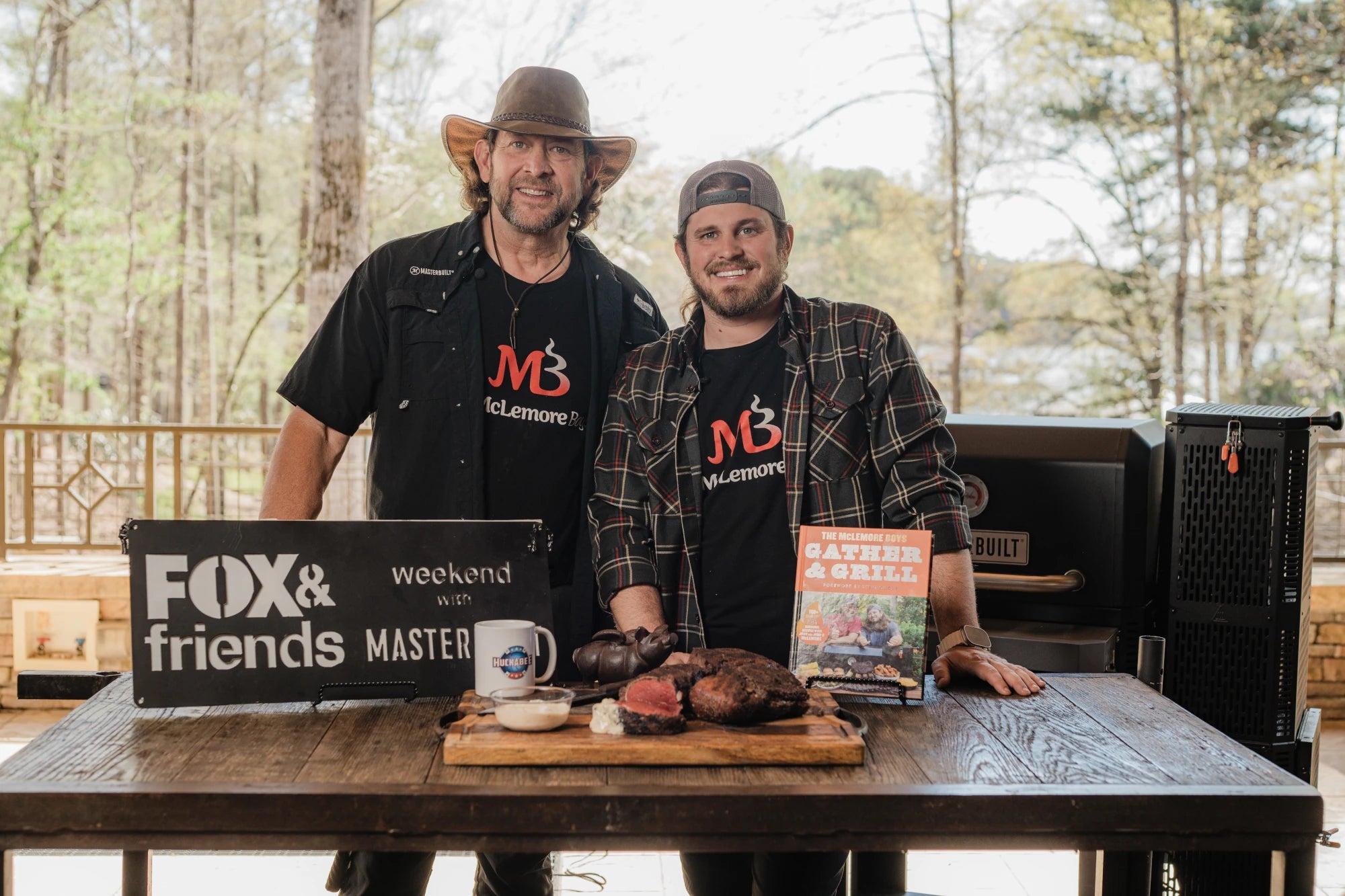 The McLemore Boys, John & John, stand next to each other behind a wooden table on an outdoor deck. On the table are a Fox & Friend weekend with Masterbuilt sign and a copy of their cookbook 