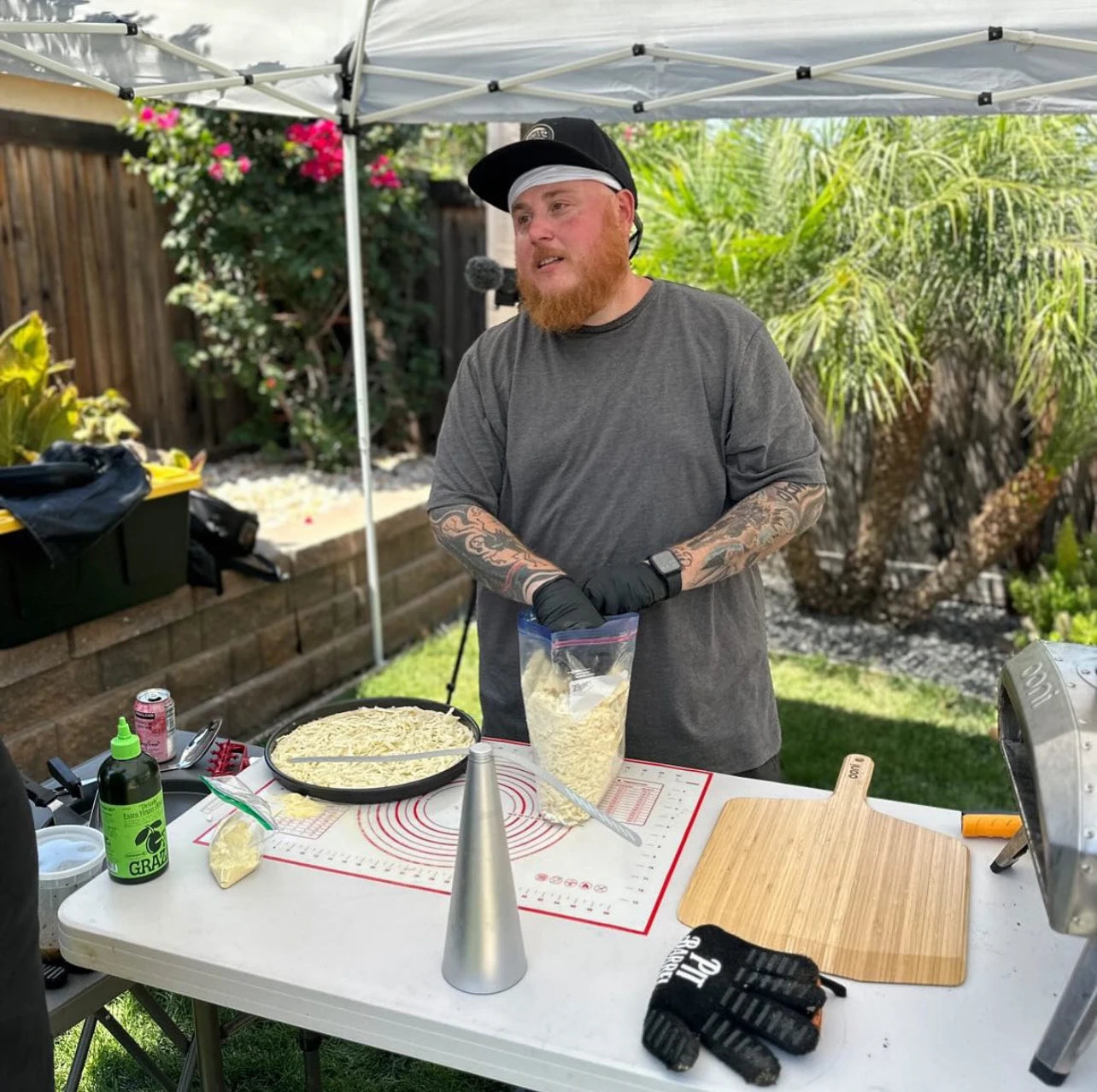 Nick Keith standing behind a table under an outdoor canopy. The table holds a wooden pizza peel, tabletop pizza oven, and other cooking accessories. 