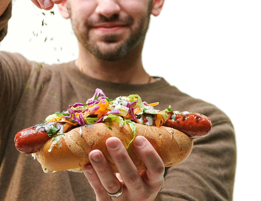 A smiling man holds a grilled sausage in a bun in one hand while he sprinkles spices on it using the other.