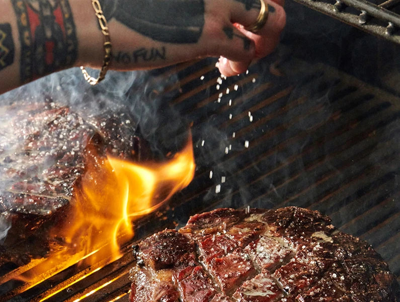 Closeup of a person with tattoos on hand and arm sprinkles salt onto steaks searing over flames coming up through a cooking grate.