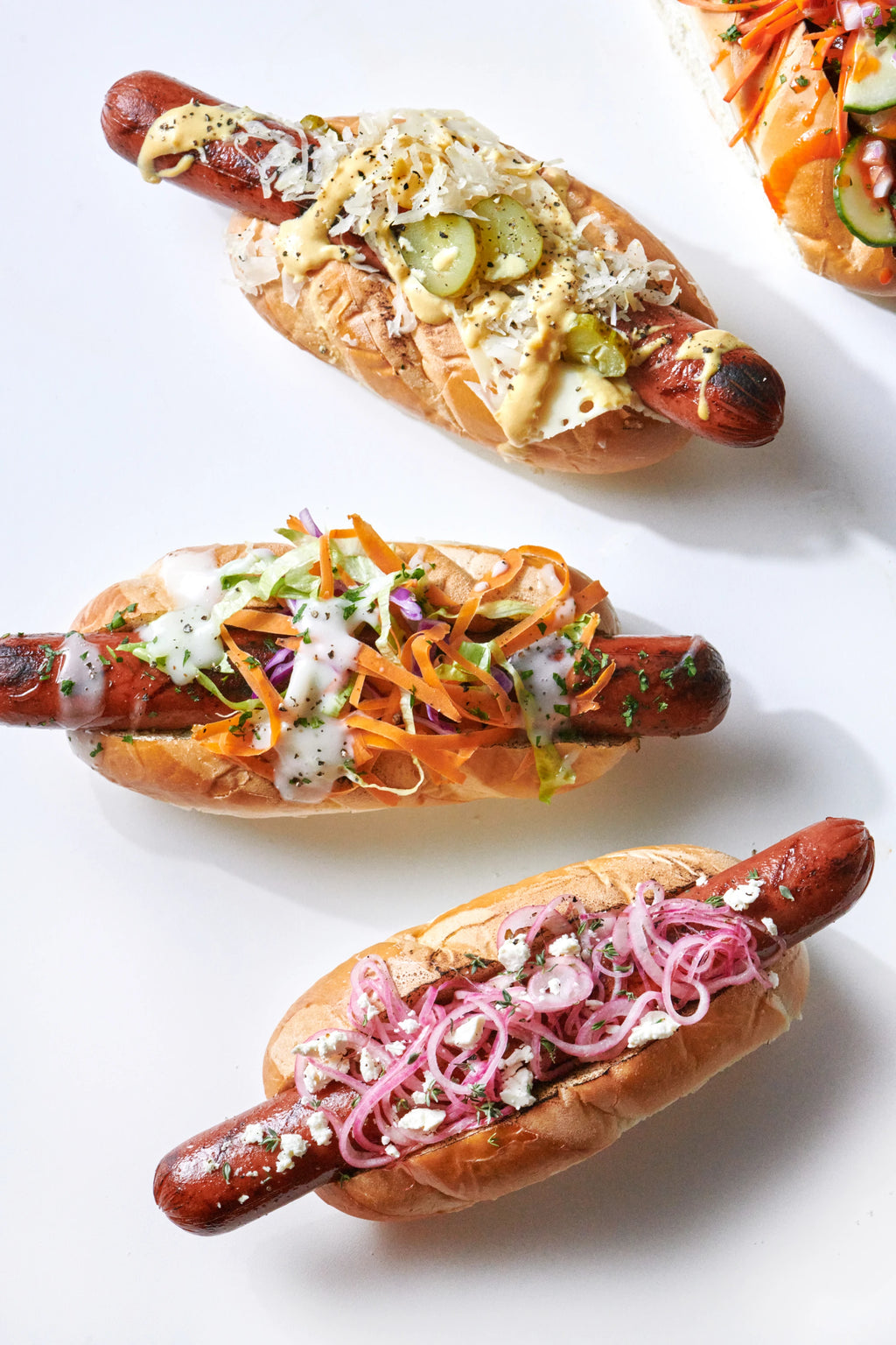 A 4 grilled sausages in buns with a variety of toppings arranged in an arc on a white background