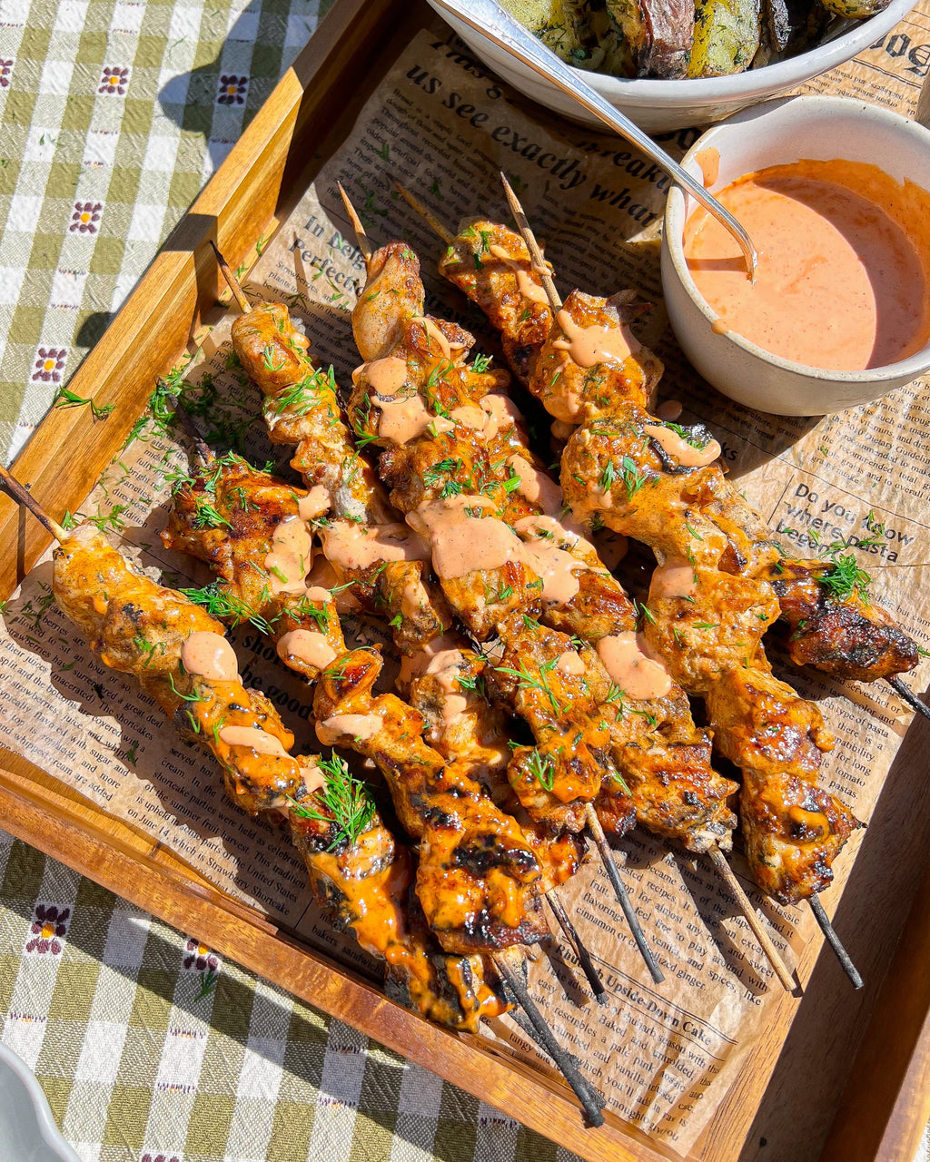 Cooked chicken skewers drizzled with Bang Bang sauce sit piled in a wooden tray lined with newspaper.