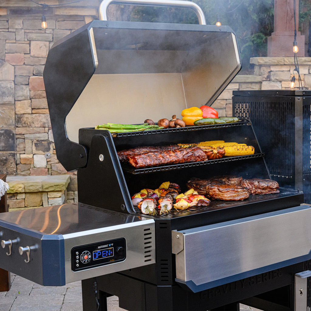 An open Gravity Series 1050 showing ribs, steaks, kebabs, winter squash halves, corn on the cob, asparagus spears, whole mushrooms, whole bell peppers, and whole chili peppers cooking on the grates and warming rack.