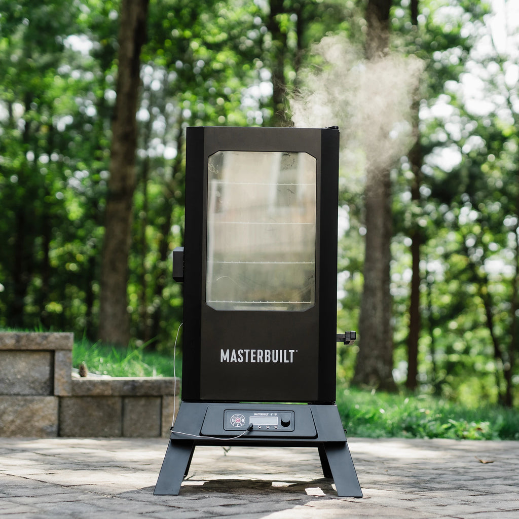 Smoke billows from the top vent of a Masterbuilt vertical smoker. 4 racks of food can barely be seen through the window in the door as smoke billows around it. A temperature probe is plugged into the digital control panel at the base of the smoker.