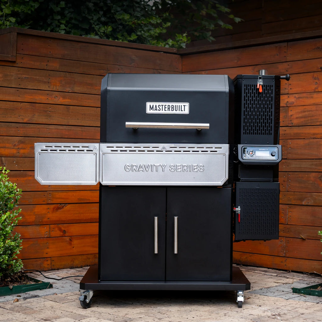 The Gravity Serie 1150 Digital Grill and Smoker on an outdoor patio in front of a wooden privacy fence. The grill sits on an enclosed cart with 4 wheels. The folding front and left side shelves are in the lowered position. The hopper and digital control panel are mounted on the right of the grill. 