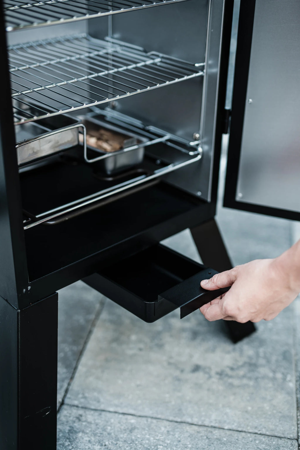 A person pulls the drip tray out from the bottom of a vertical smoker