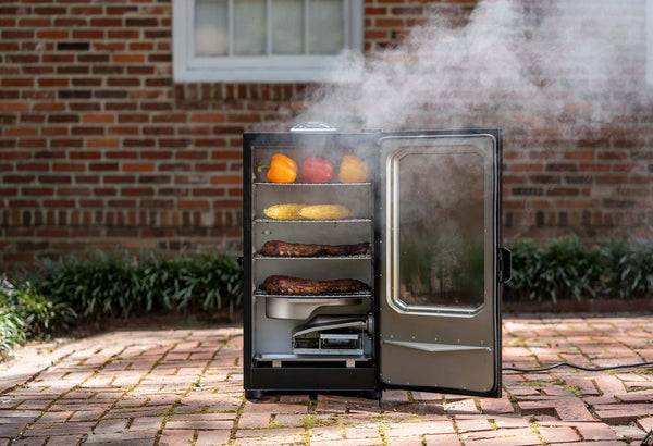 Smoke billows out of an open smoker with  4 racks full of food. The removeable water bowl and wood chip loader system are below the cooking racks.