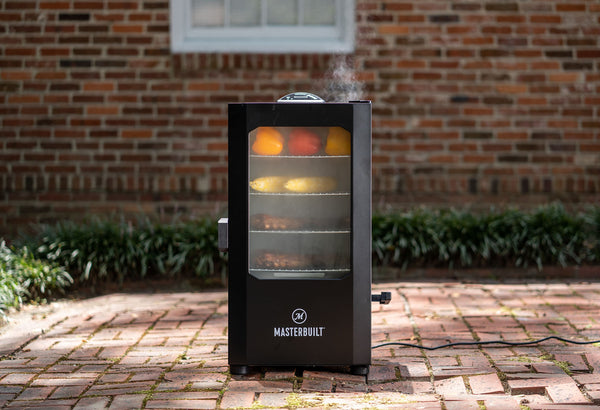 A smoker sits on a brick patio in front of a brick house with a white window. The smoker holds bell peppers, corn on the cob, and 2 racks of ribs.